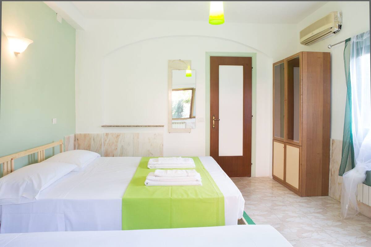 A hotel room with two beds, a green bedspread, a wooden wardrobe, a door, and an air conditioning unit.