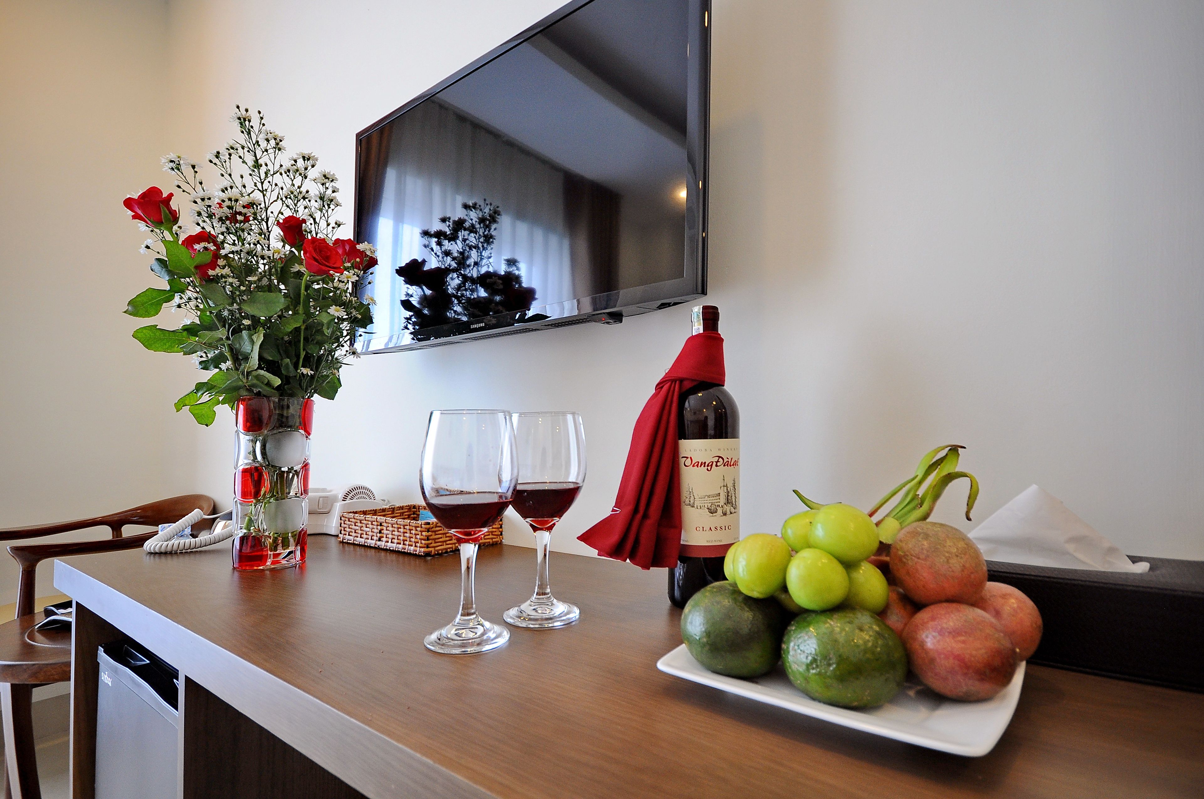 Una mesa con un jarrón de rosas rojas, dos copas de vino, una botella de vino y un plato de fruta.
