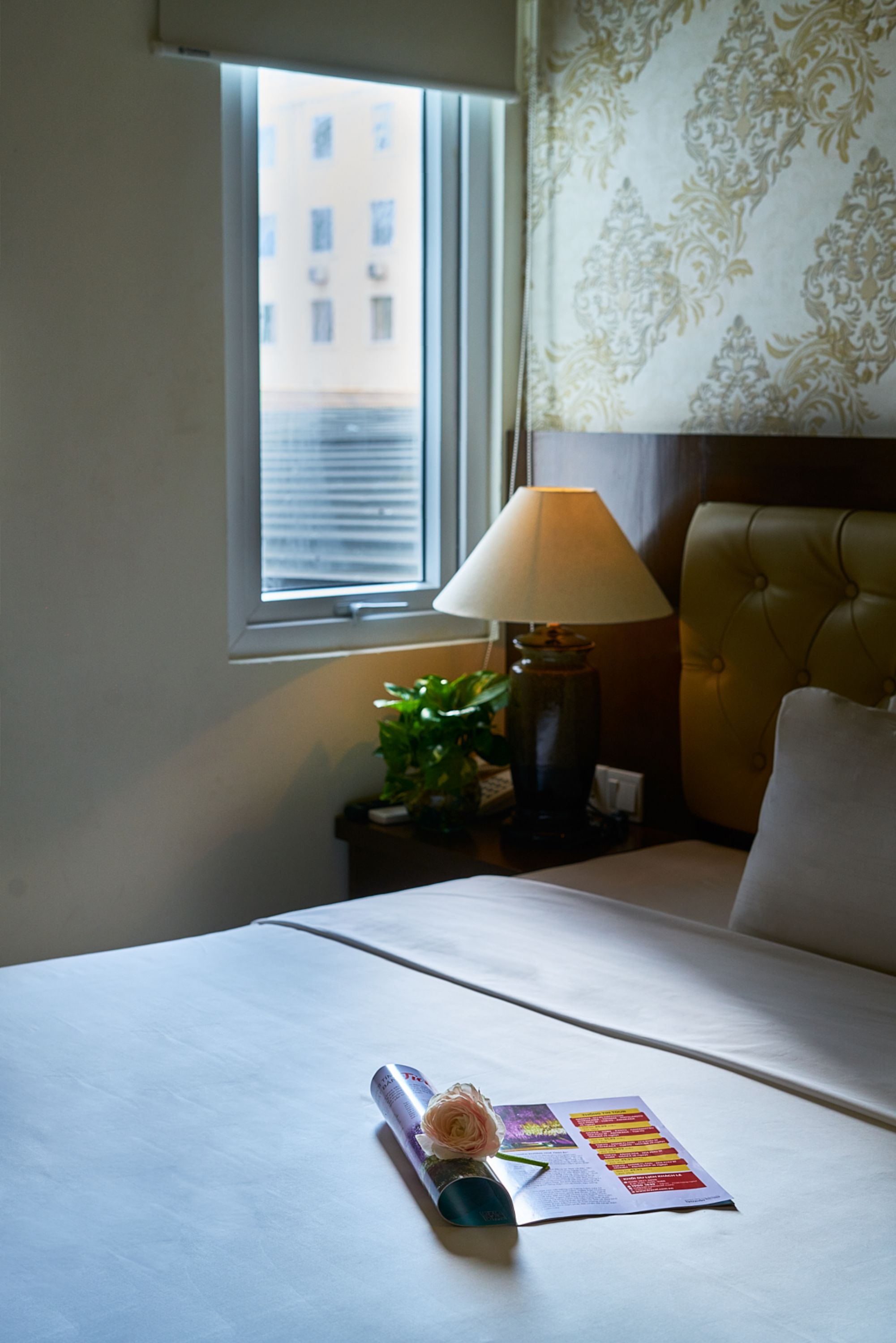 A hotel room with a bed, a nightstand with a lamp, a window with a view of buildings, and a decorative wall pattern.