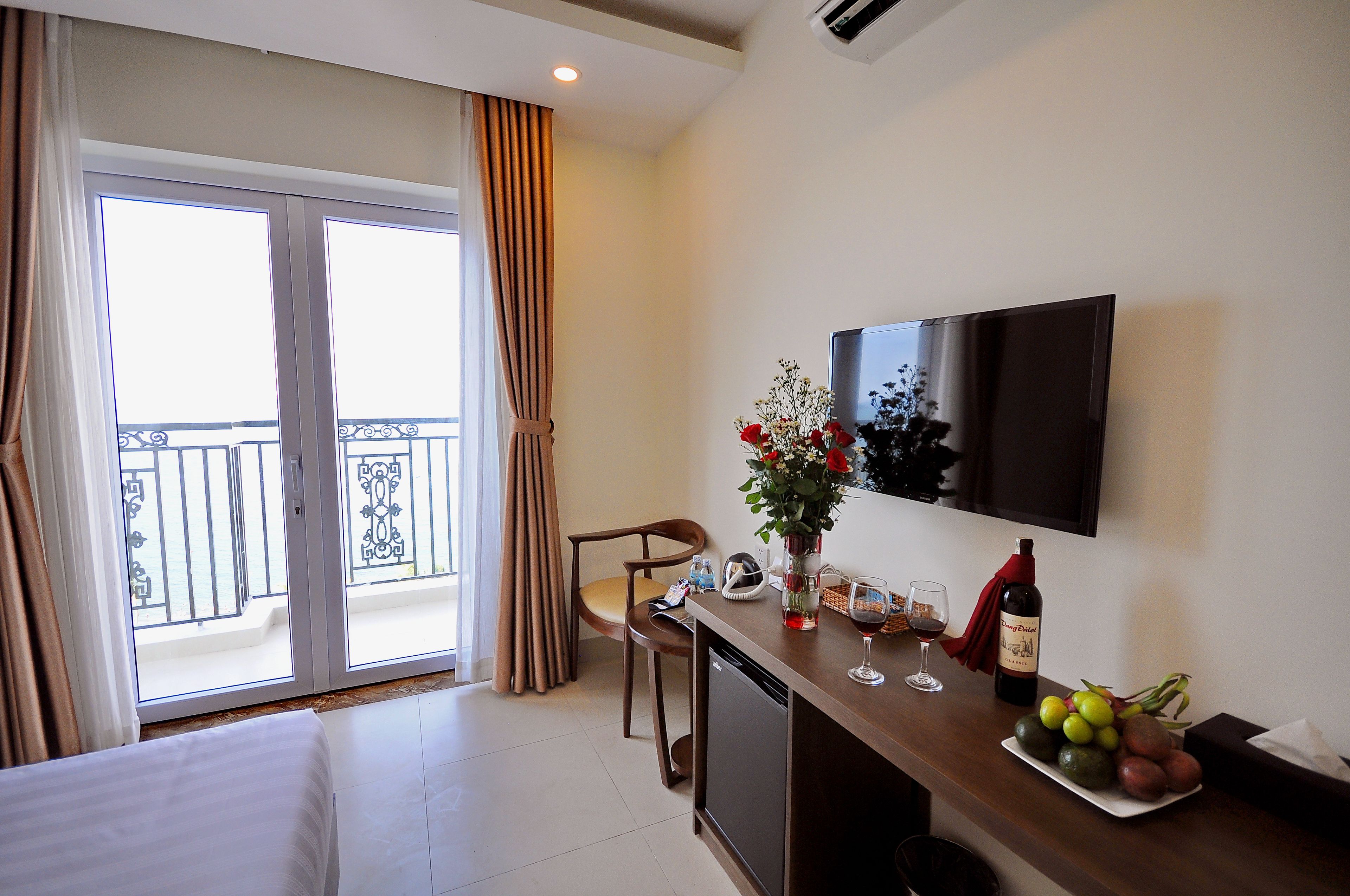 A hotel room with a balcony, a dining area with a table and chairs, a TV mounted on the wall, and a small refrigerator.