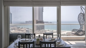 Una mesa de comedor dispuesta con platos, vasos y cubiertos, con vista a un paisaje urbano costero con rascacielos y una playa.