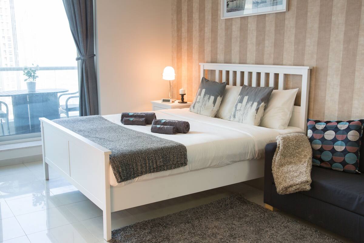 Un dormitorio moderno con cama blanca, almohadas grises y blancas, una lámpara de noche, un sofá oscuro y vistas al exterior.