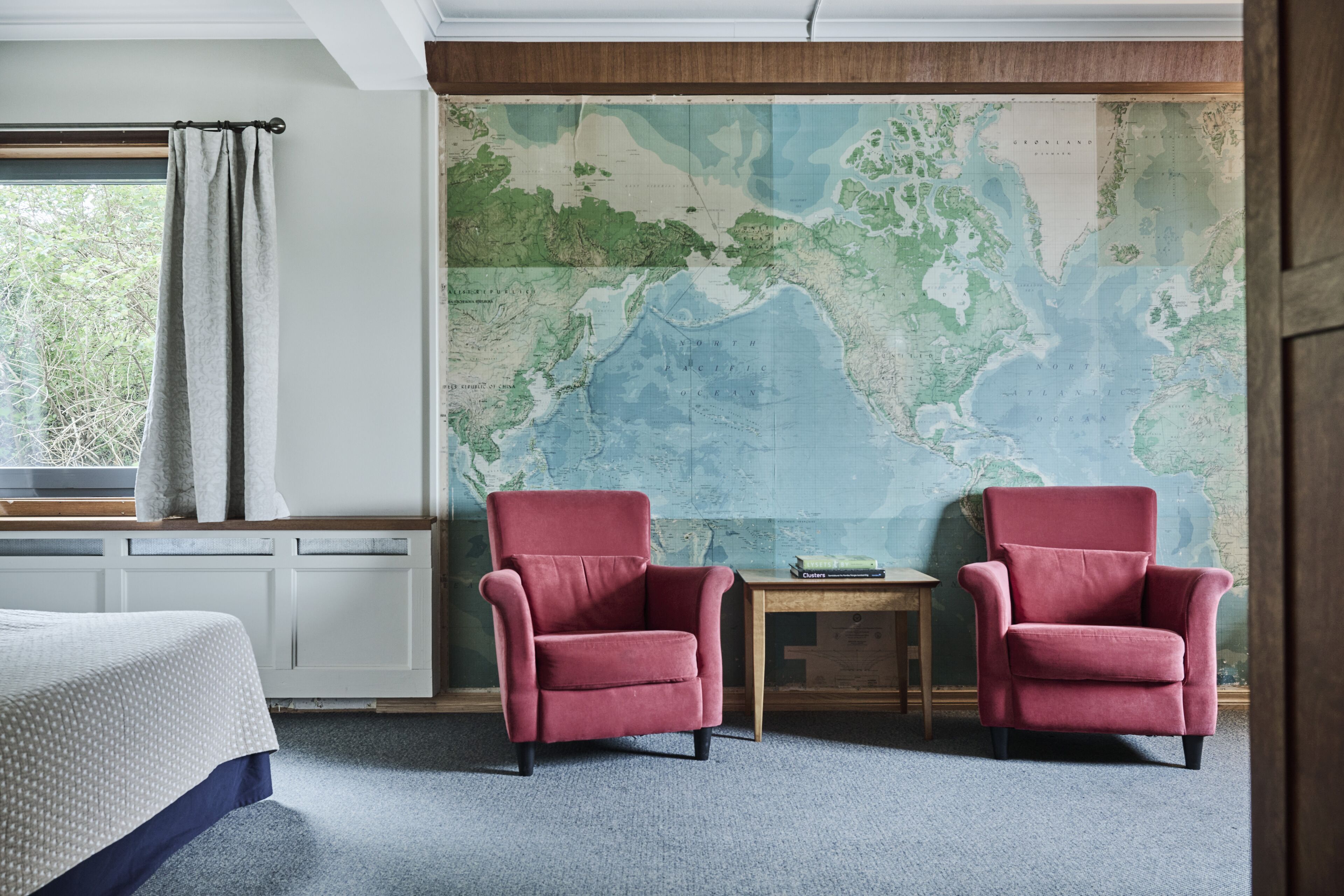 A room with a large map on the wall, two red armchairs, a wooden table, and a bed with a white and blue bedspread.