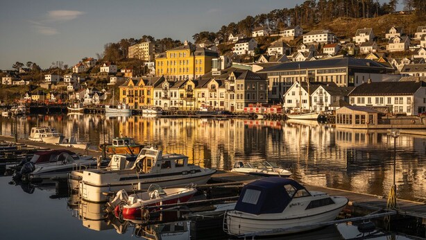 A harbor with boats docked, buildings with varied architecture, and a hillside in the background. - Rederiet Hotell (Farsund)