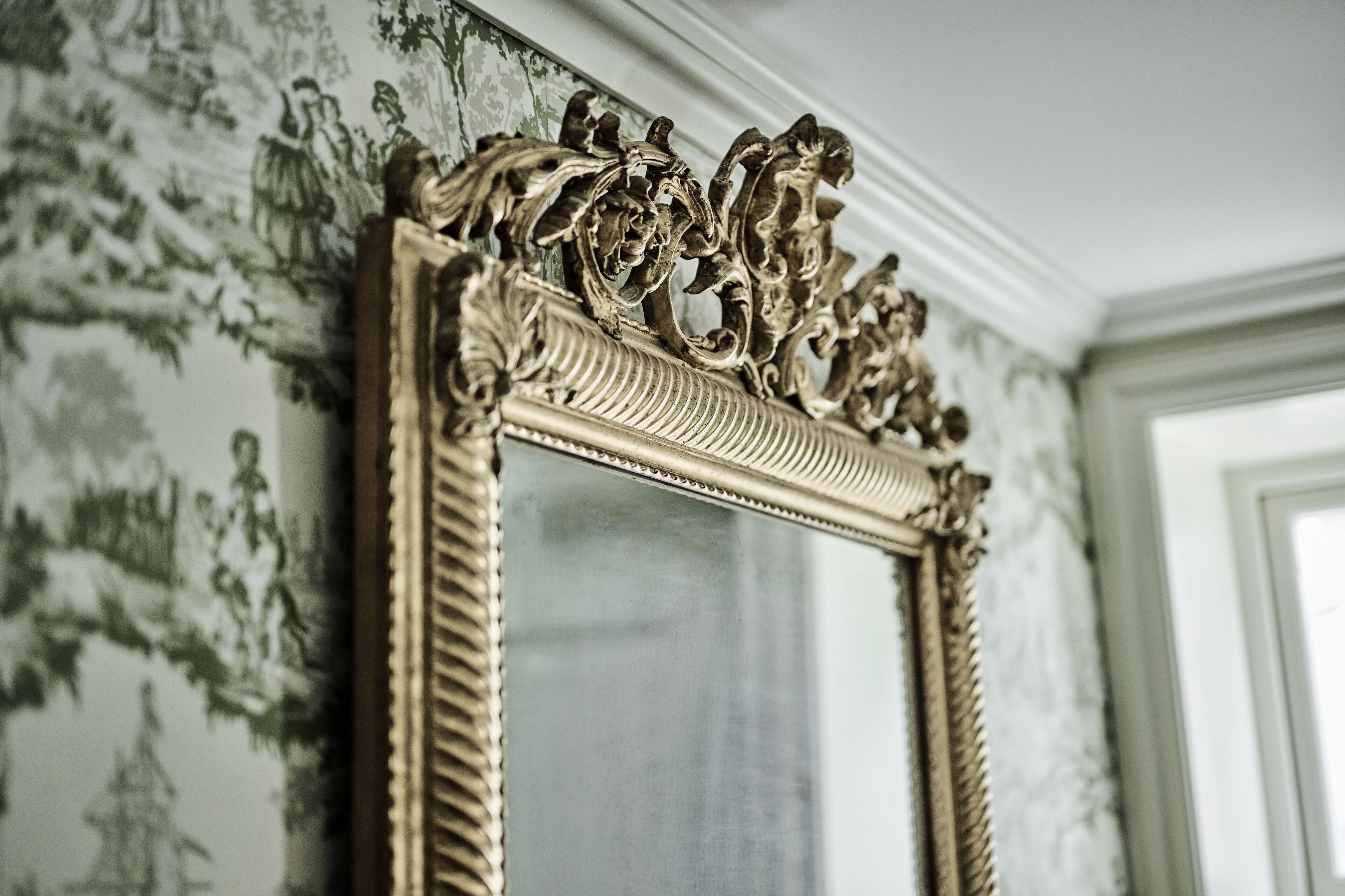 A detailed ornate mirror with a decorative frame, set against a wallpaper with a floral pattern.