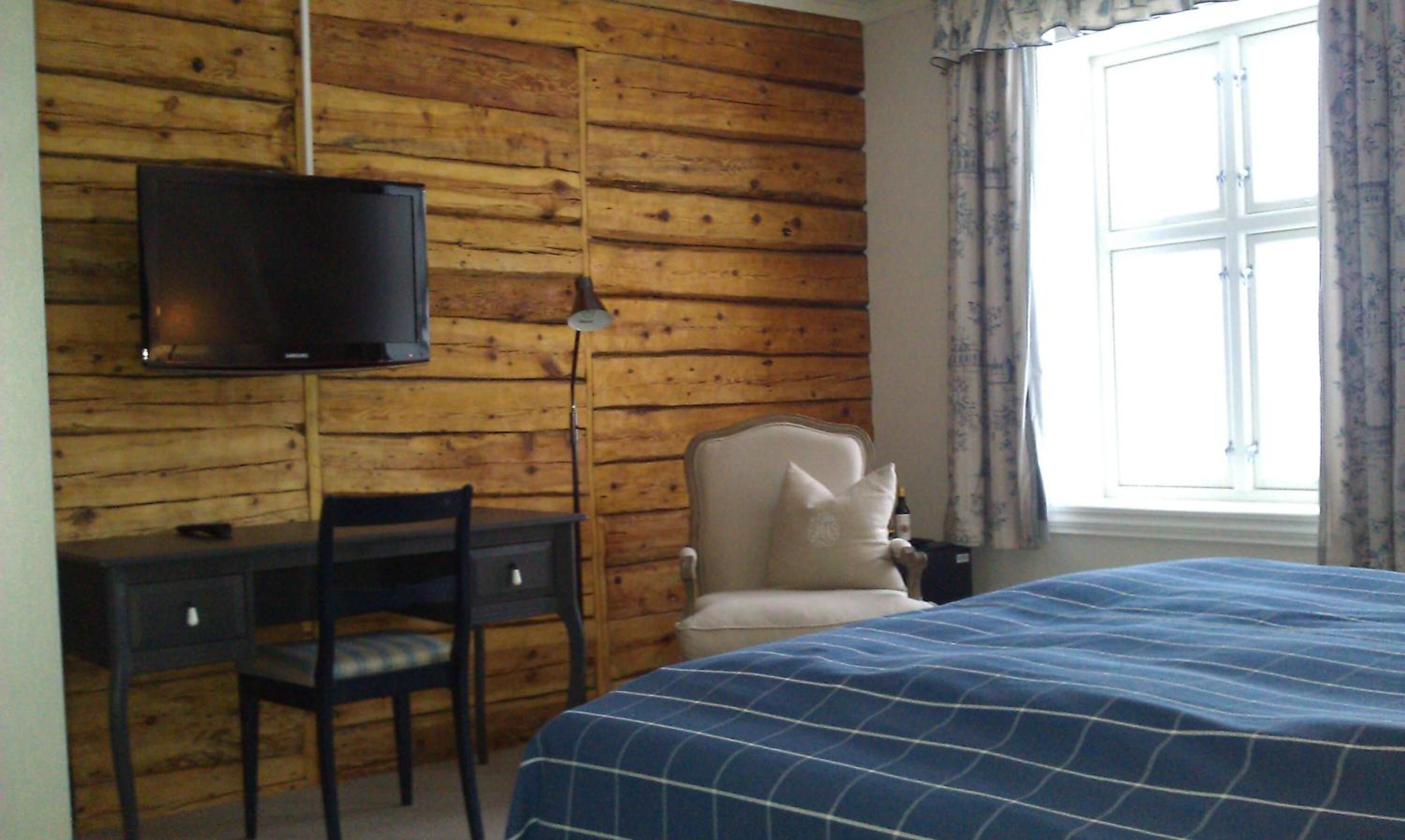 Una habitación con pared de madera, una cama con cubrecama azul, un escritorio con silla y un televisor montado en la pared.