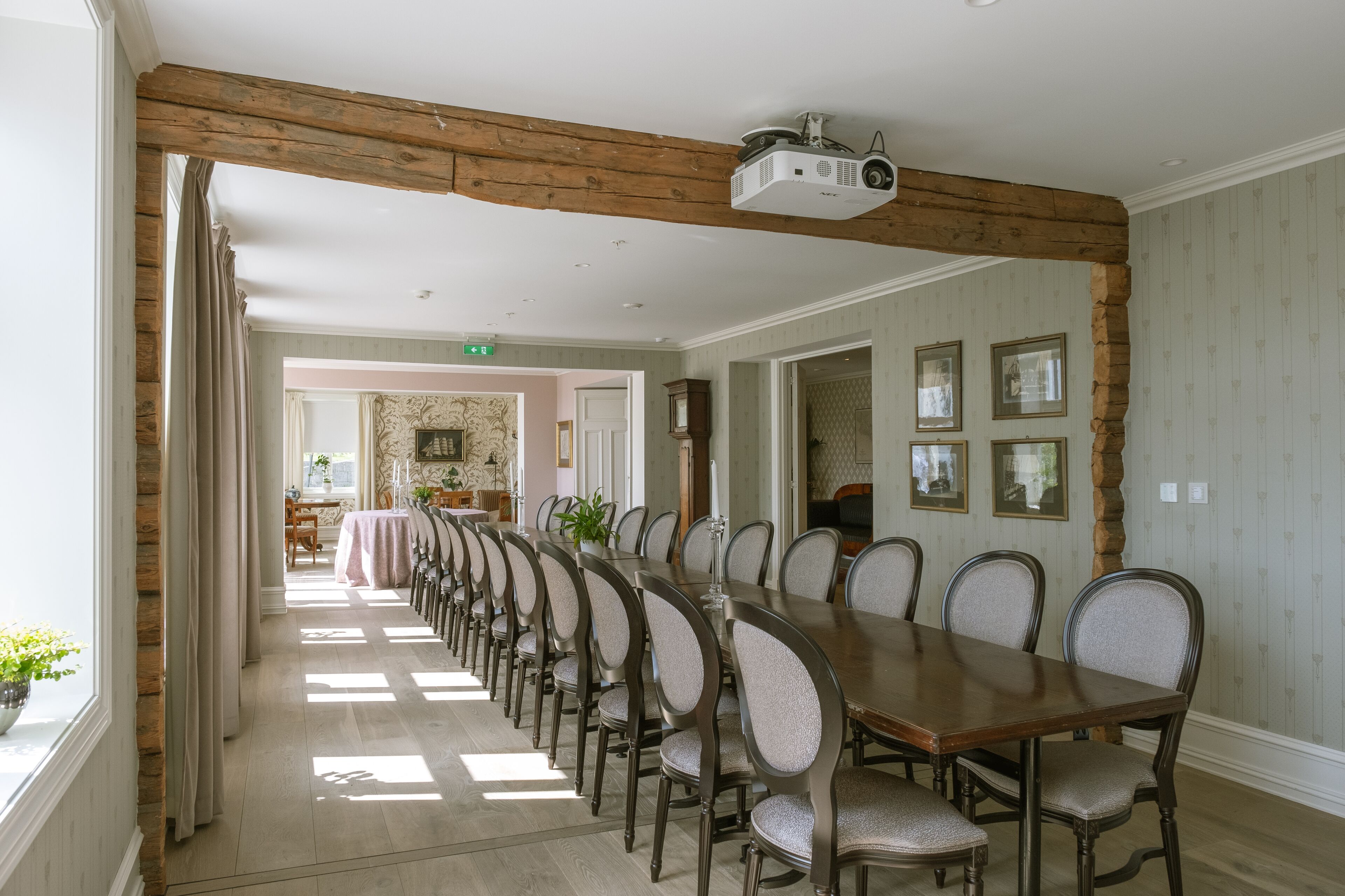 Een lange eettafel met stoelen, houten balken aan het plafond en ingekaderde foto's aan de muur.