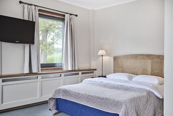 A hotel room with a bed, a flat-screen TV mounted on the wall, a window with curtains, a bedside lamp, and a view of greenery outside.