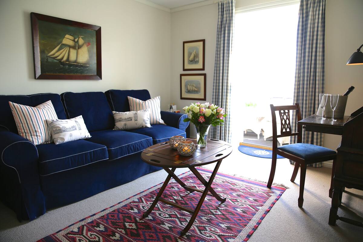 A living room with a blue sofa, a patterned rug, a small table with a vase of flowers, and a chair.