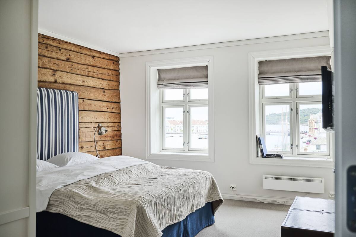 A bedroom with a wooden accent wall, a bed with a striped pillow, two windows with curtains, and a view of buildings outside.