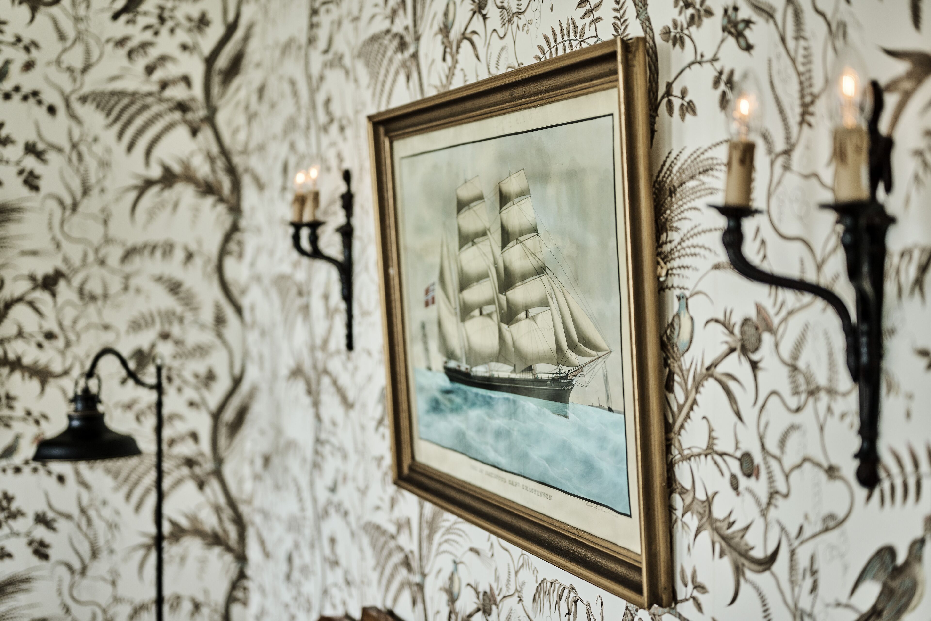 A framed maritime painting on a wall with ornate wallpaper, a wall-mounted candelabra, and a floor lamp.