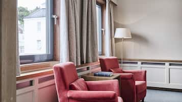 Una habitación con dos sillones rojos, una mesita con un libro, una lámpara de pie y un ventanal con cortinas.