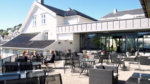 An outdoor patio area with tables and chairs, a swimming pool, and a building with a sloped roof in the background. - Rederiet Hotell (Farsund)