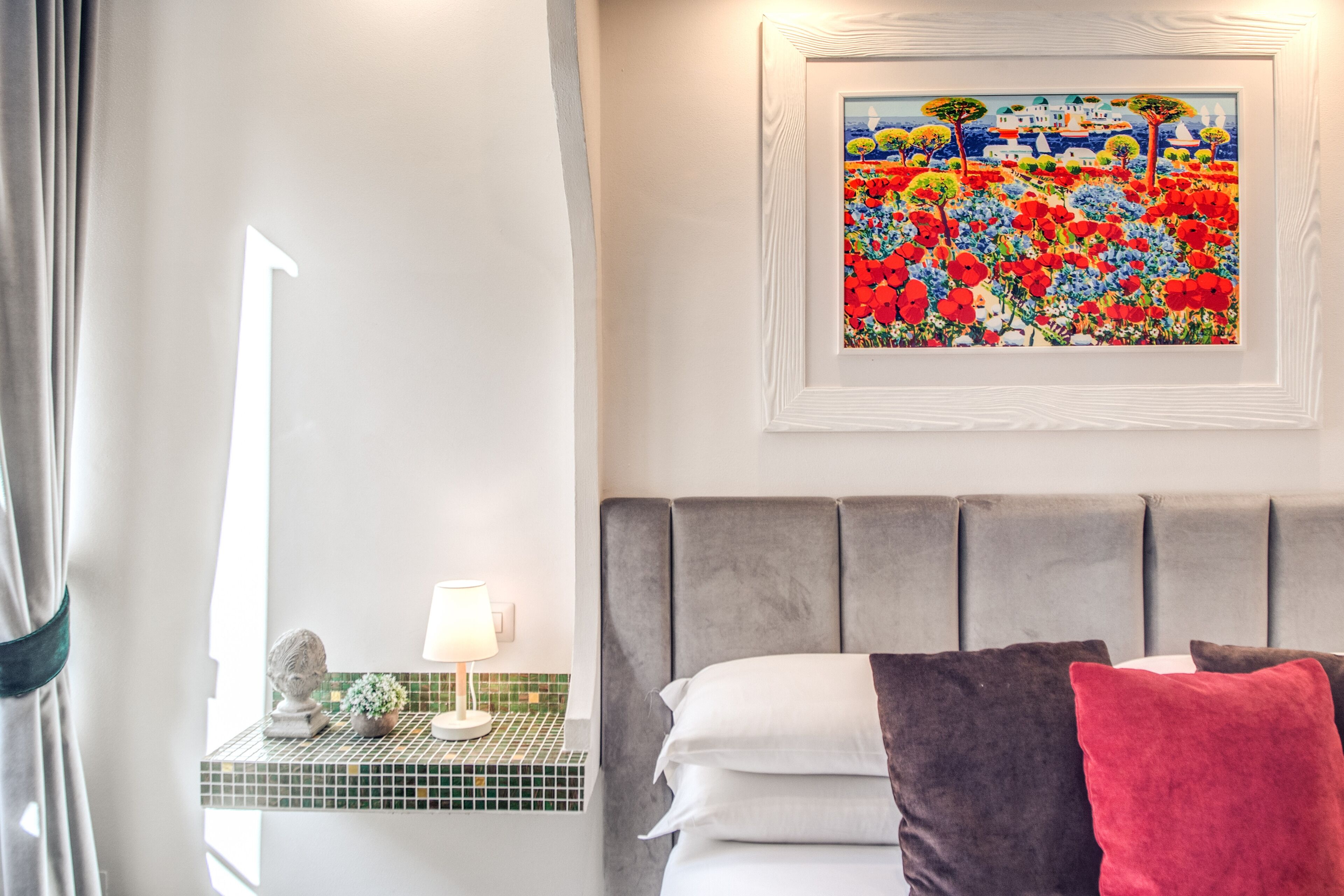 A hotel room with a bed, a nightstand with a lamp, a framed painting, and a view of the sea.