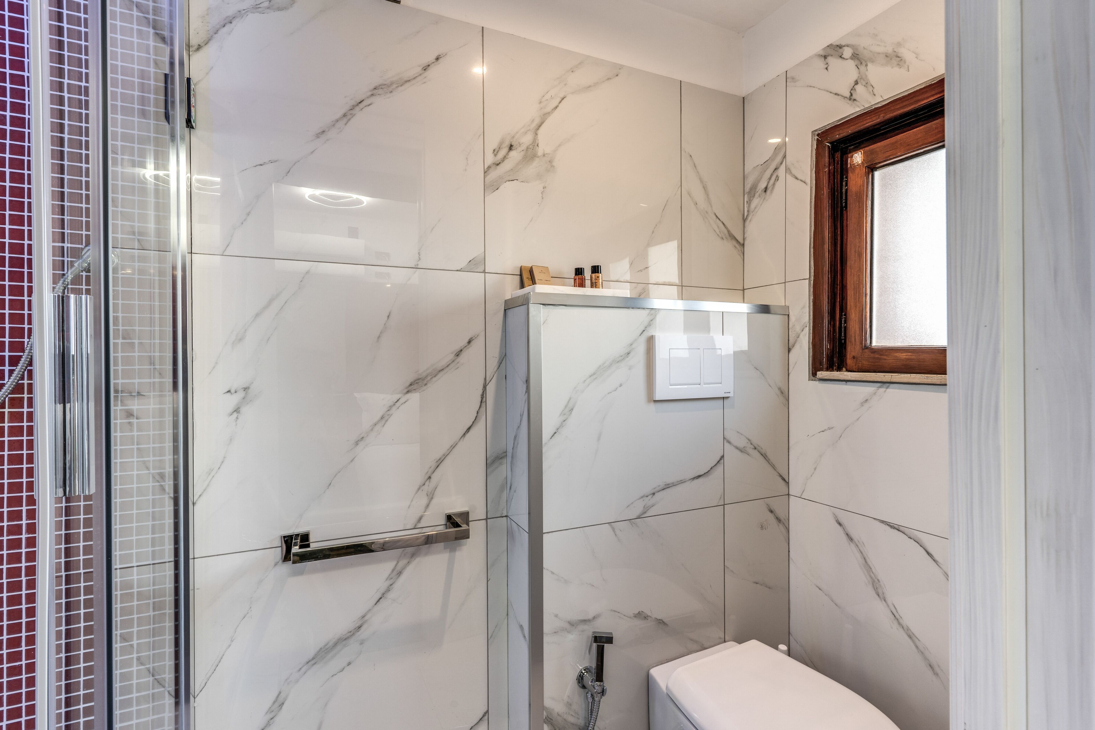 A modern bathroom with marble walls, a glass shower enclosure, and a wooden-framed window.