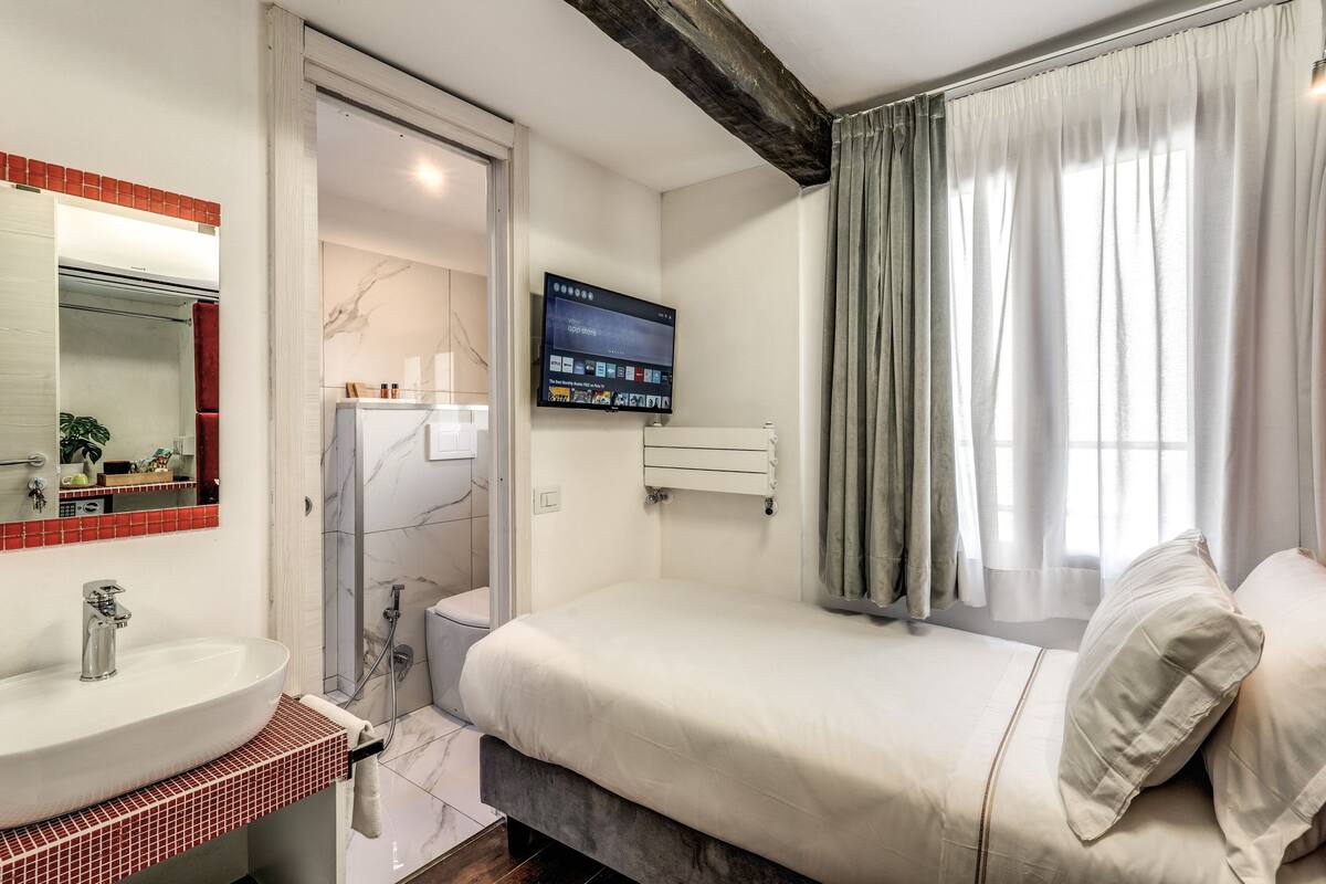 A modern bedroom with a large bed, a built-in TV, a bathroom with a shower, and a red-tiled sink area.
