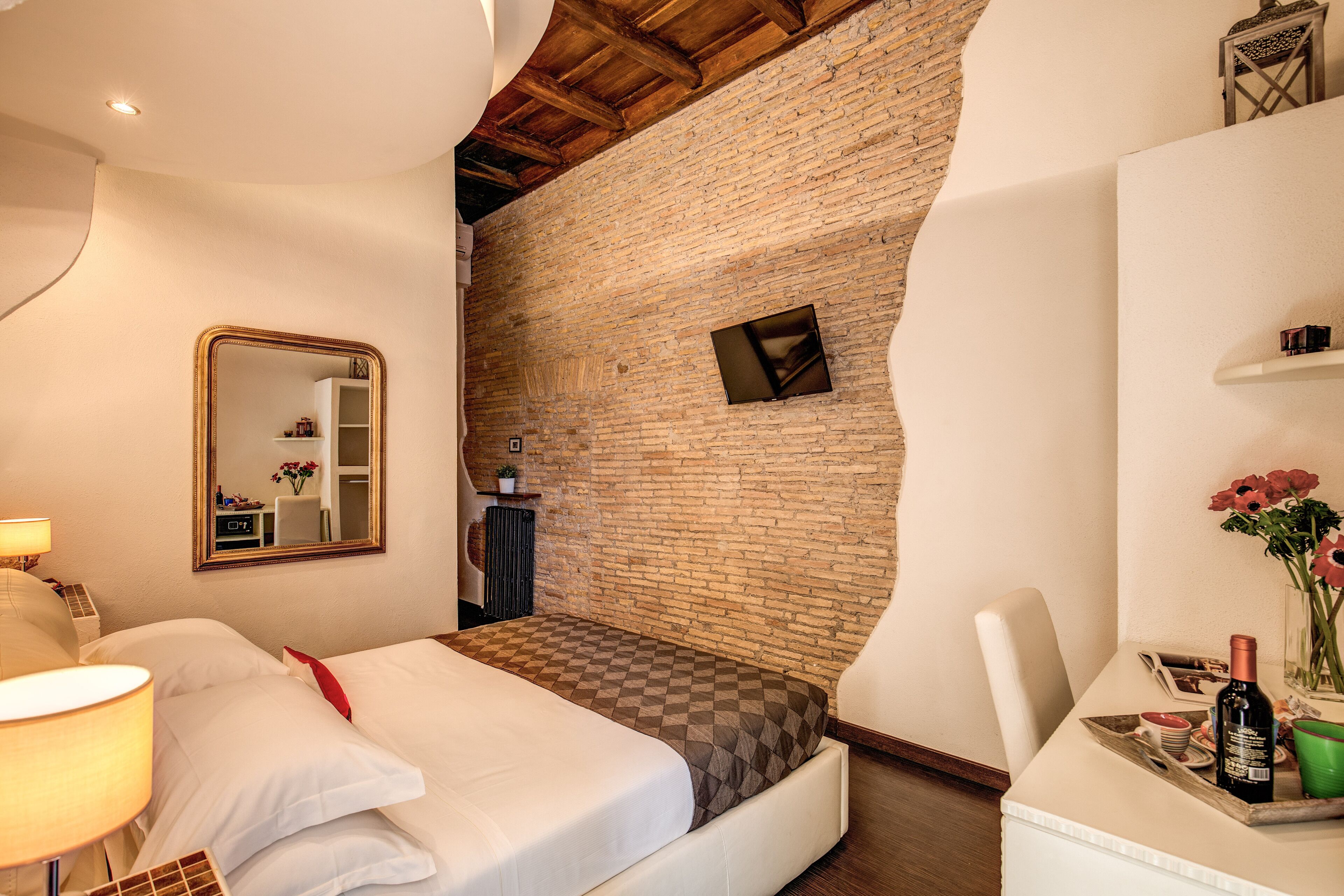 A hotel room with a checkered bed, a large mirror, a brick wall, and a small table with a bottle and flowers.