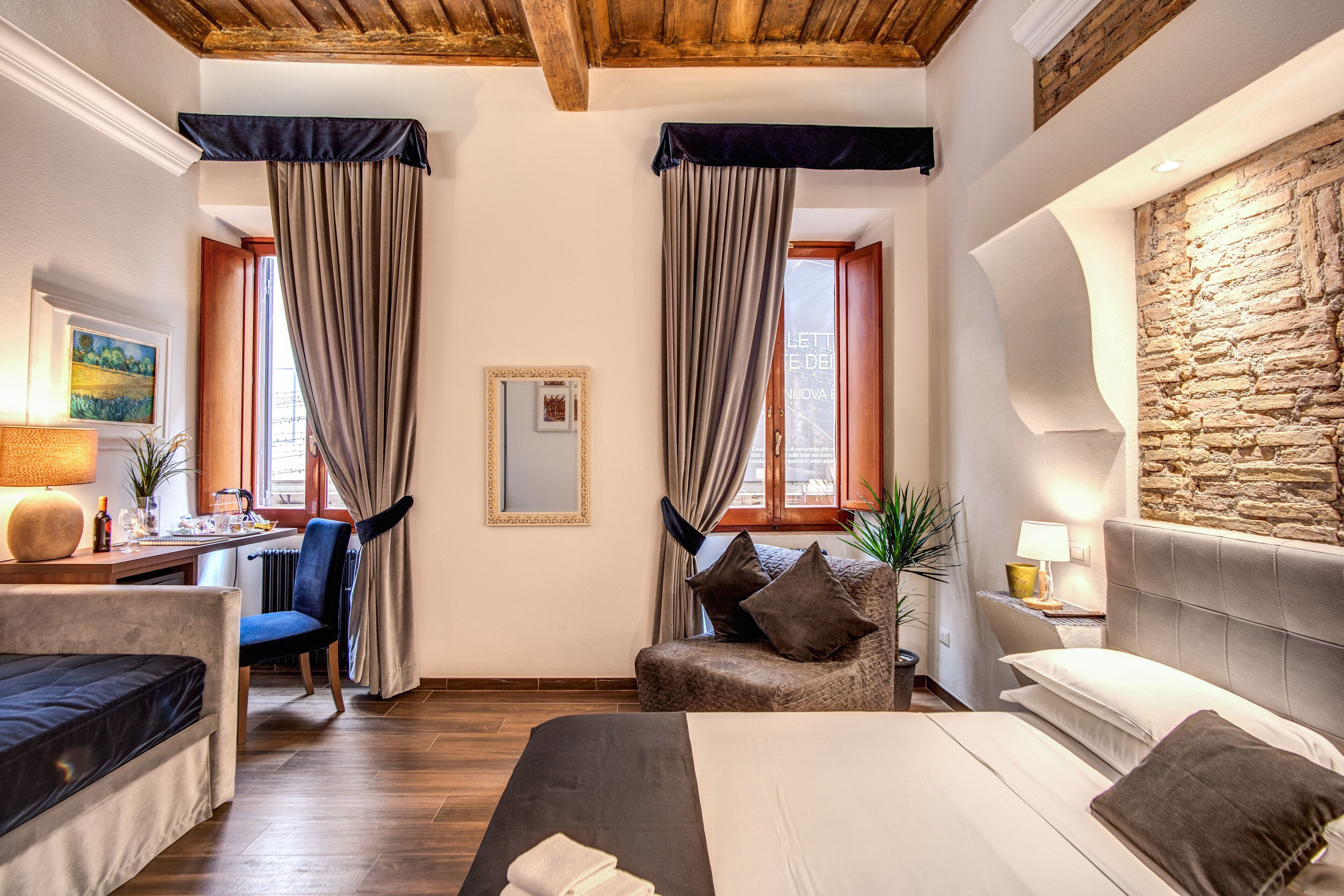 Une chambre d’hôtel avec un lit, un bureau, une chaise et un canapé. Il y a des rideaux aux fenêtres, un tableau au mur et un mur en pierre sur un des côtés.