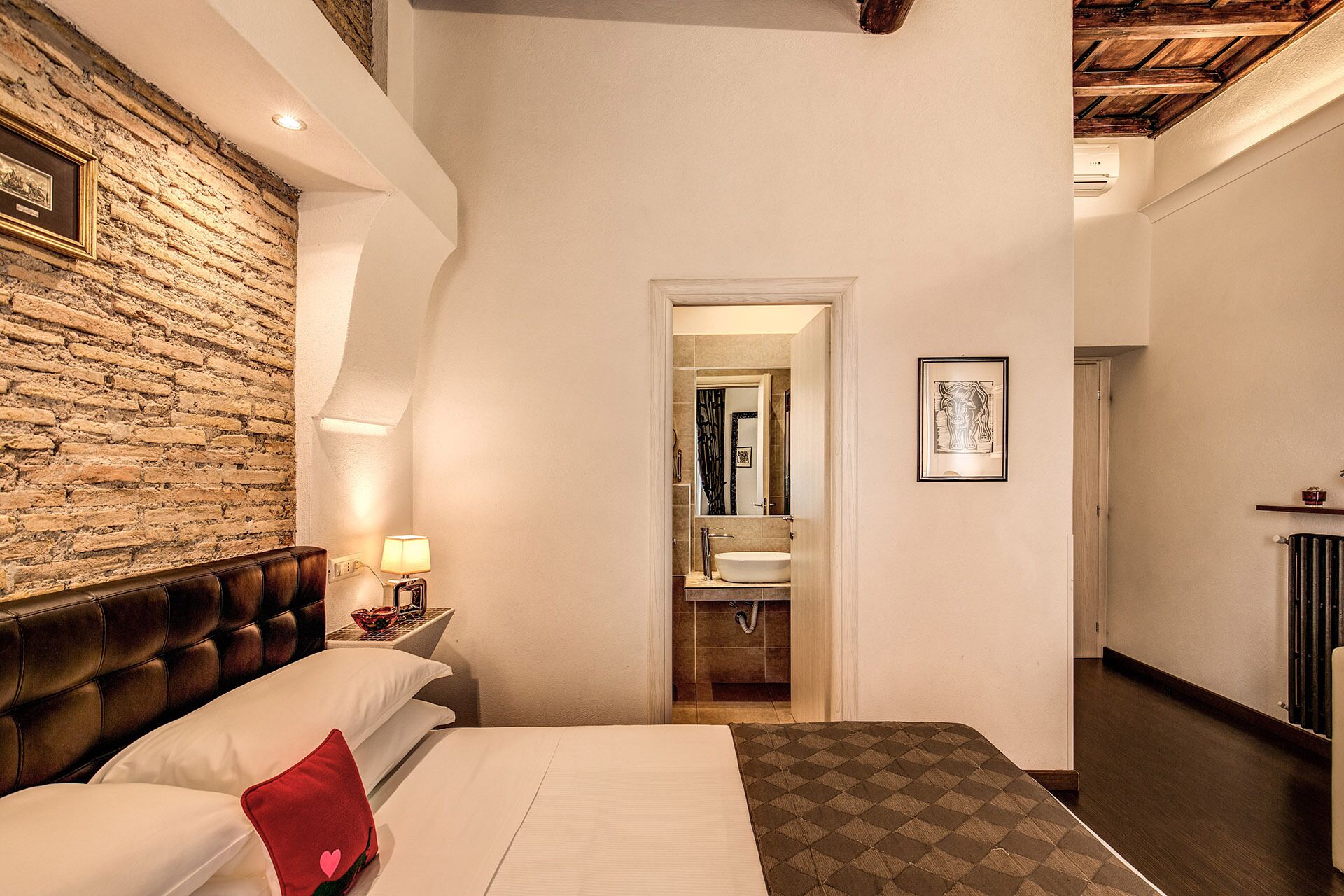 A hotel room with a bed, a nightstand, a lamp, a brick wall, a mirror, and a door leading to a bathroom.