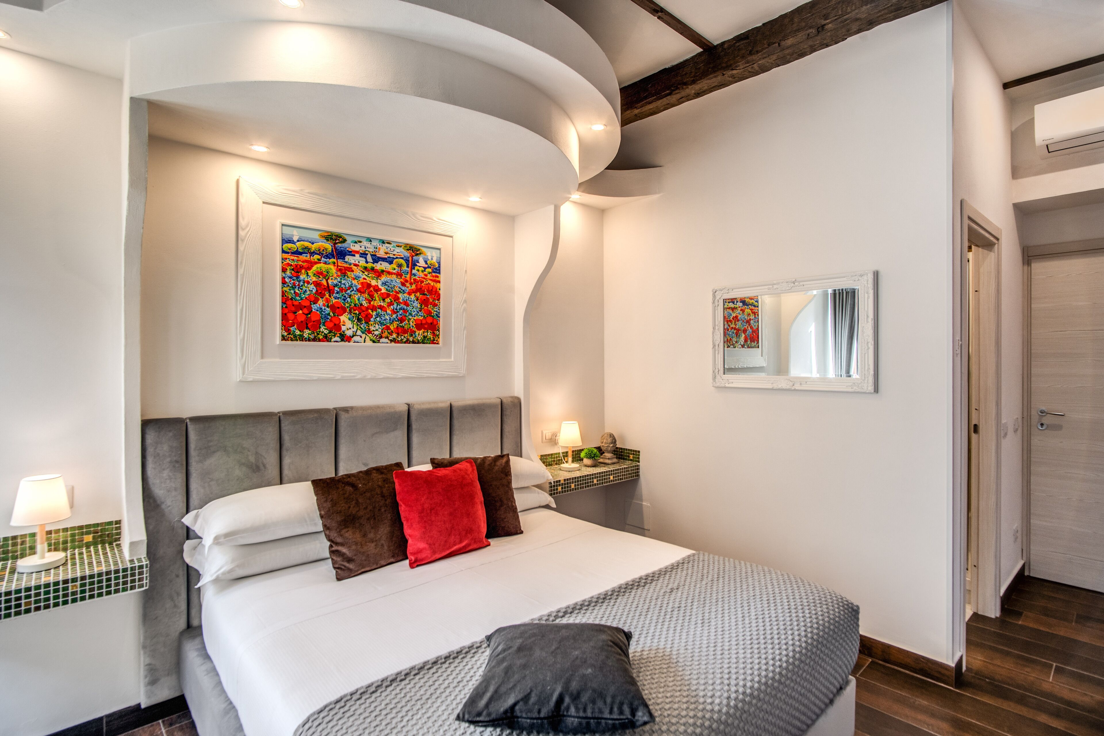 A modern bedroom with a large bed, a headboard, a nightstand, a lamp, a painting, and a mirror.