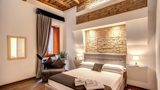 A bedroom with a stone wall headboard, a large bed with pillows and towels, a wooden ceiling, and a nightstand with a lamp. - Piazza di Spagna Comfort Rooms (Rome)