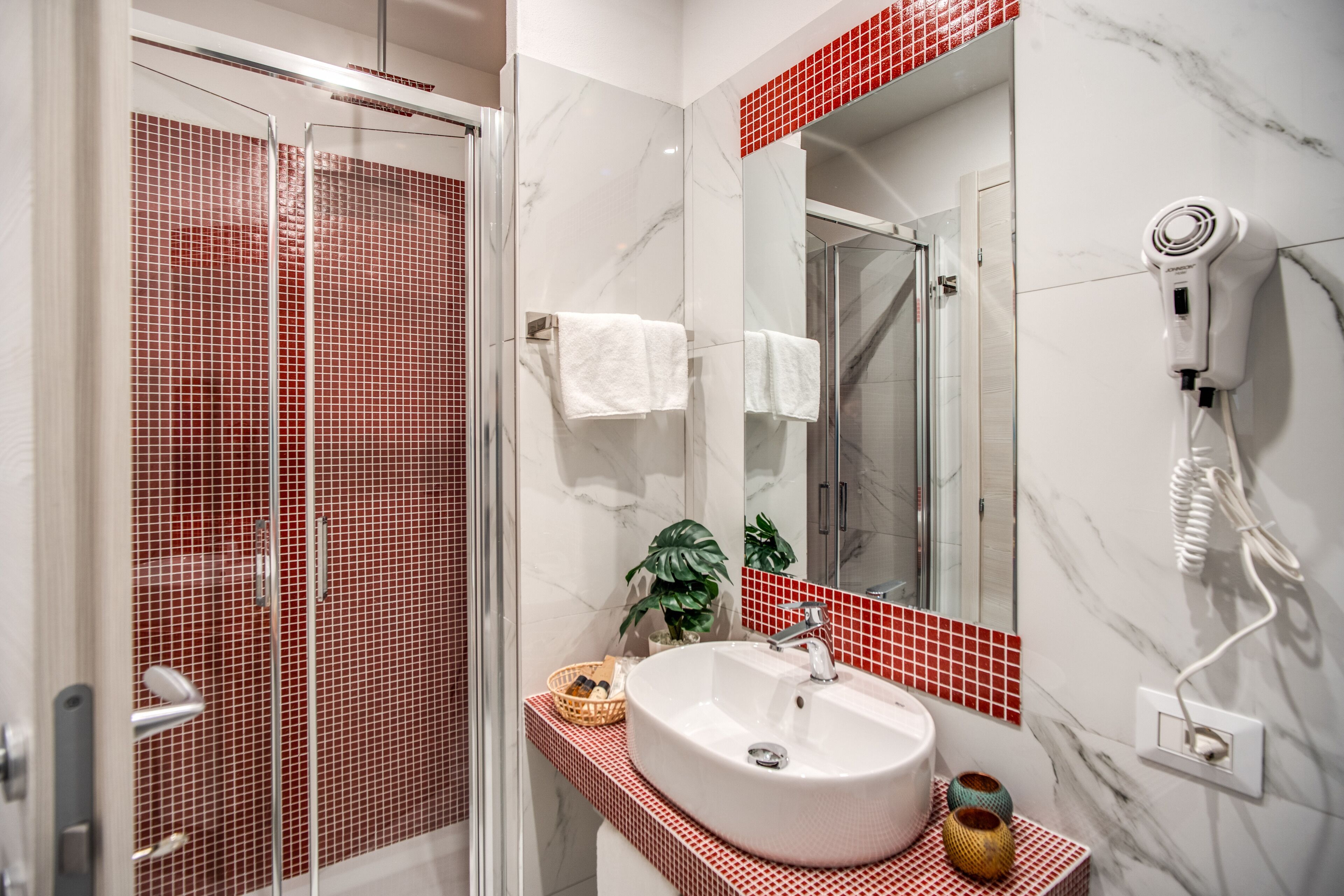 A bathroom with a glass shower enclosure, a white sink with a red-tiled countertop, and a hairdryer mounted on the wall.