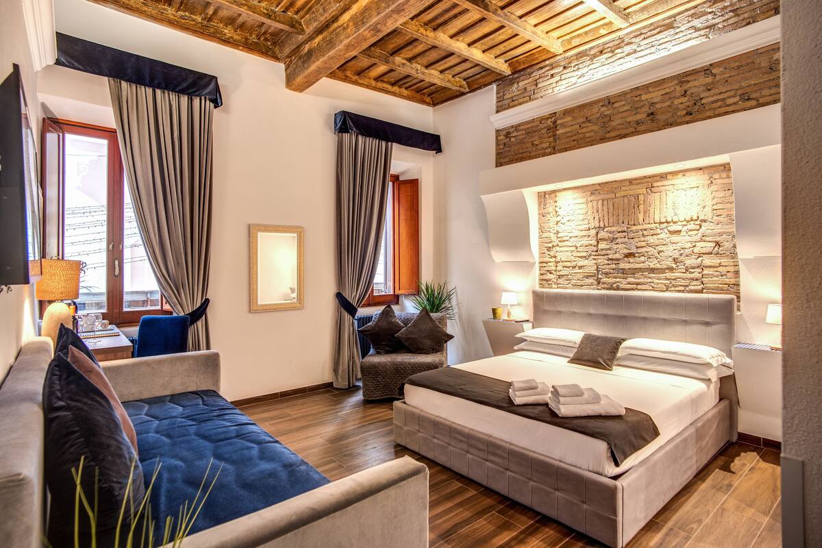 A modern hotel room with a large bed, a sofa, a dining table, and a stone accent wall.