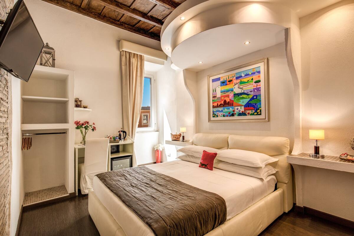 A hotel room with a bed, a desk, a TV, a painting, and a window with curtains.