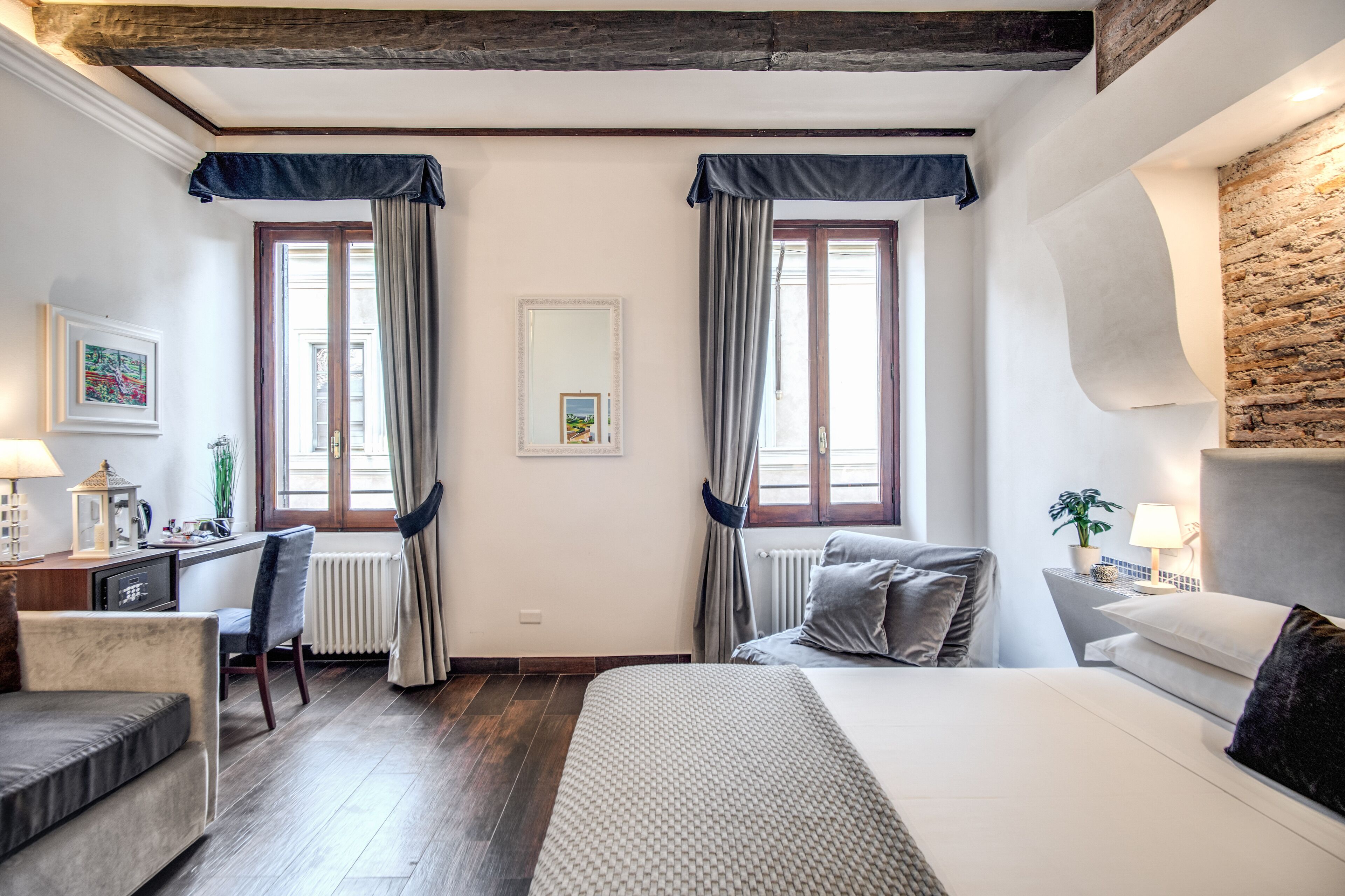A hotel room with a bed, a desk, a chair, a radiator, and two windows with curtains.