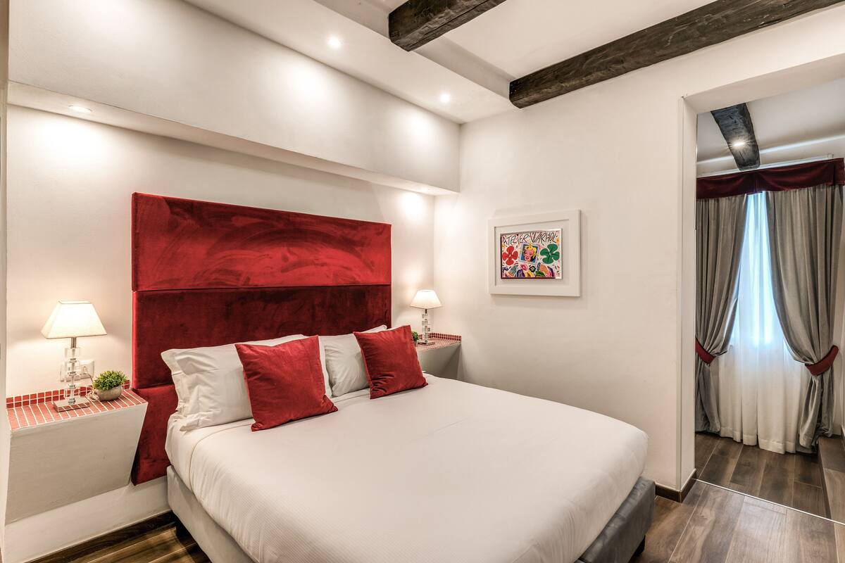 A modern hotel room with a large bed, red accent pillows, a red headboard, and a framed artwork on the wall.