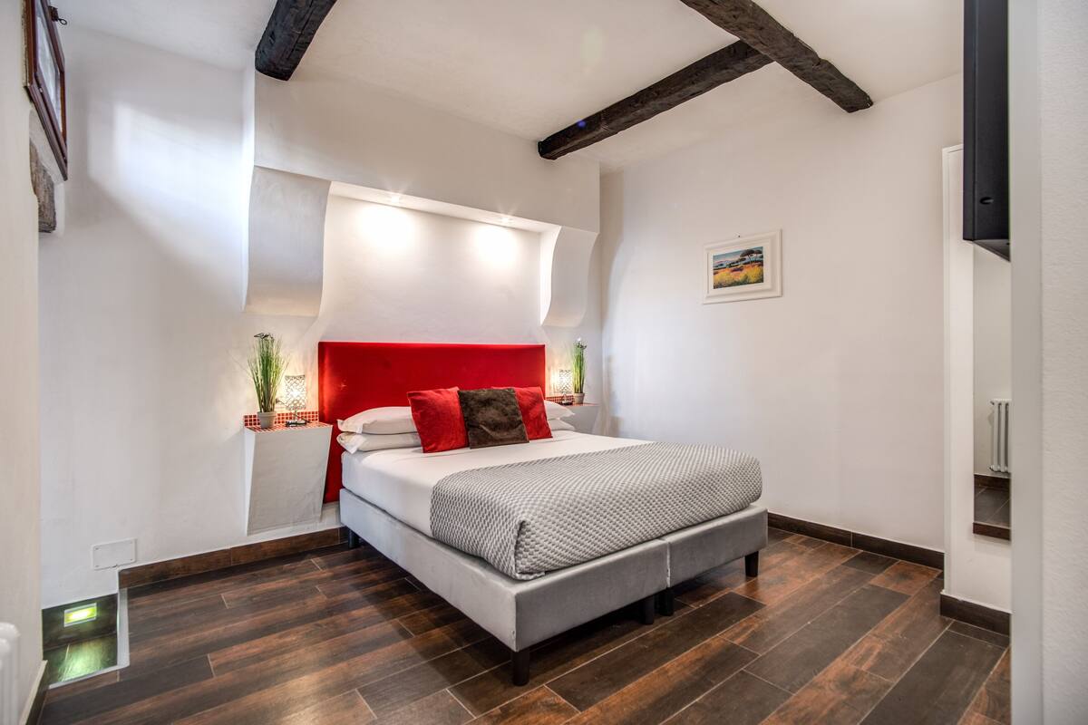 A bedroom with a bed, a red headboard, a nightstand with a lamp, and a potted plant.