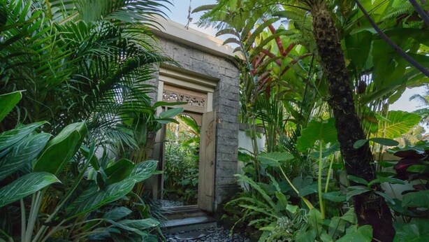 A lush garden with a stone pathway leading to a wooden door. - Jani's Place Cottage (Ubud)