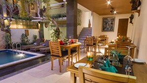 A dining area with wooden tables and chairs, a pool, and indoor plants. - Jani's Place Cottage (Ubud)