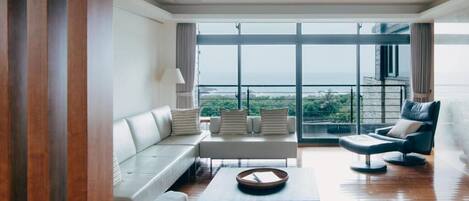 A spacious living room with a large window offering a view of the sea. There is a white sofa, a black armchair, and a coffee table.