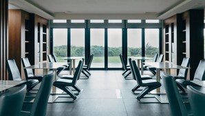 A modern dining area with tables and chairs arranged for groups, large windows offering a view of greenery, and built-in shelving units along the walls. - Adagio Shitiping (Fengbin)