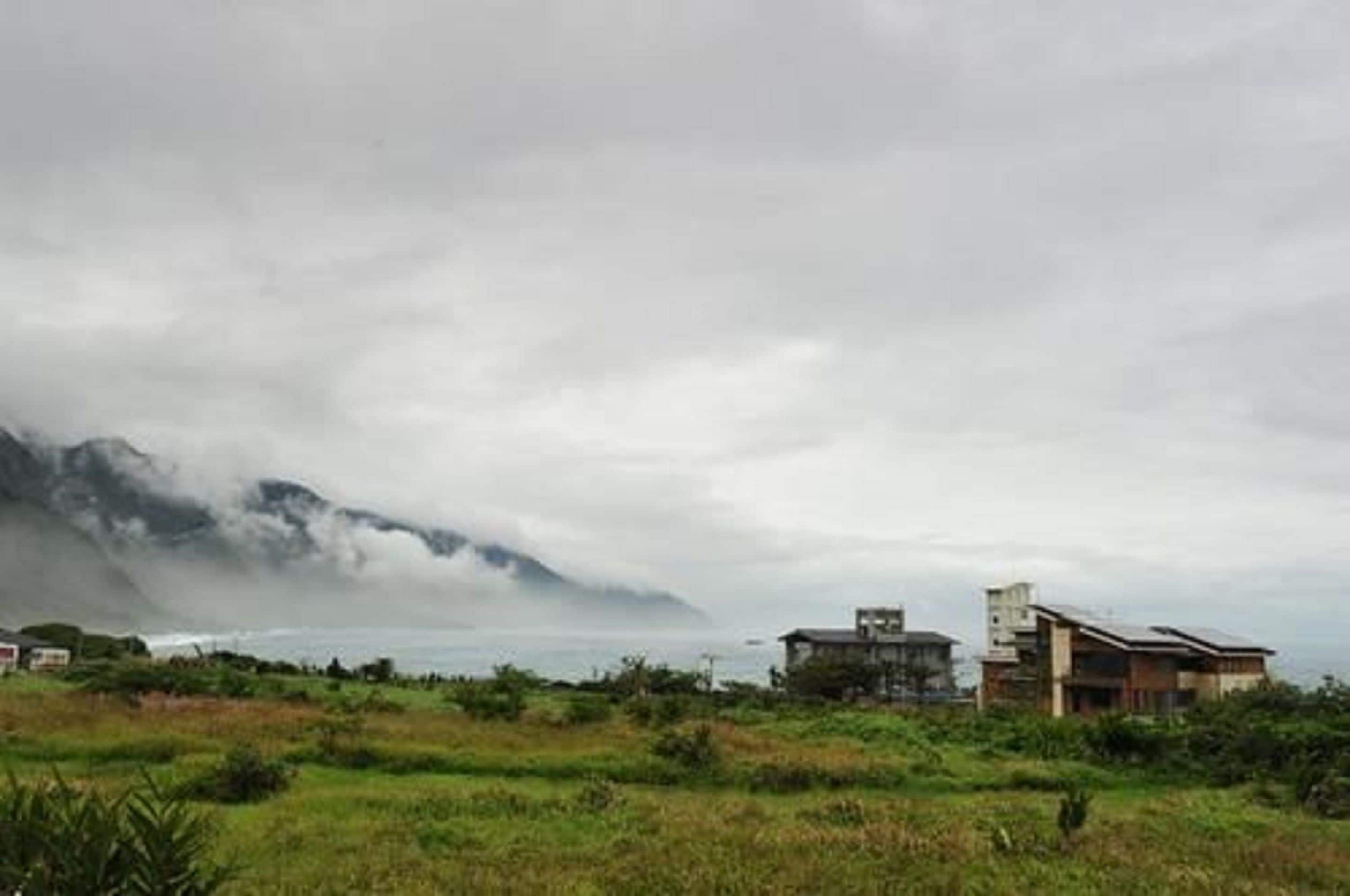 Paisagem costeira com uma montanha parcialmente coberta por nuvens, e dois edifícios com telhados planos em primeiro plano.