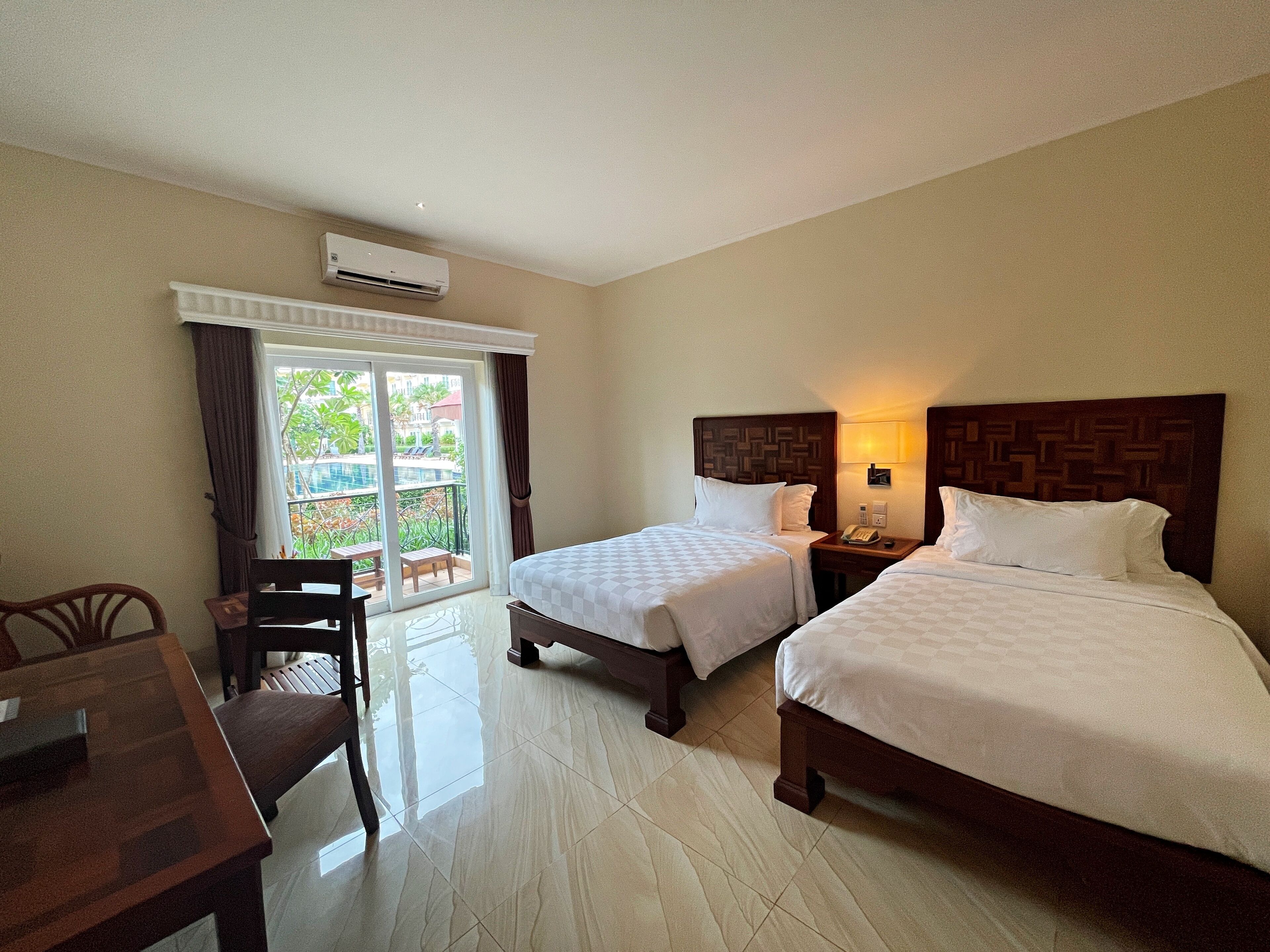 Habitación de hotel con dos camas, un escritorio y una silla. Tiene una ventana con cortinas, un equipo de aire acondicionado y vistas a una piscina y vegetación.
