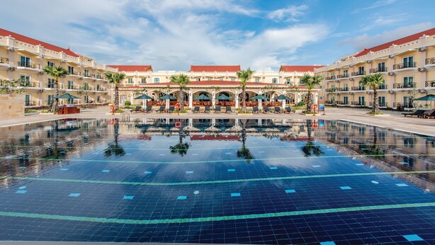 Una gran piscina con agua cristalina, rodeada por un hotel con múltiples balcones y zonas de estar al aire libre.