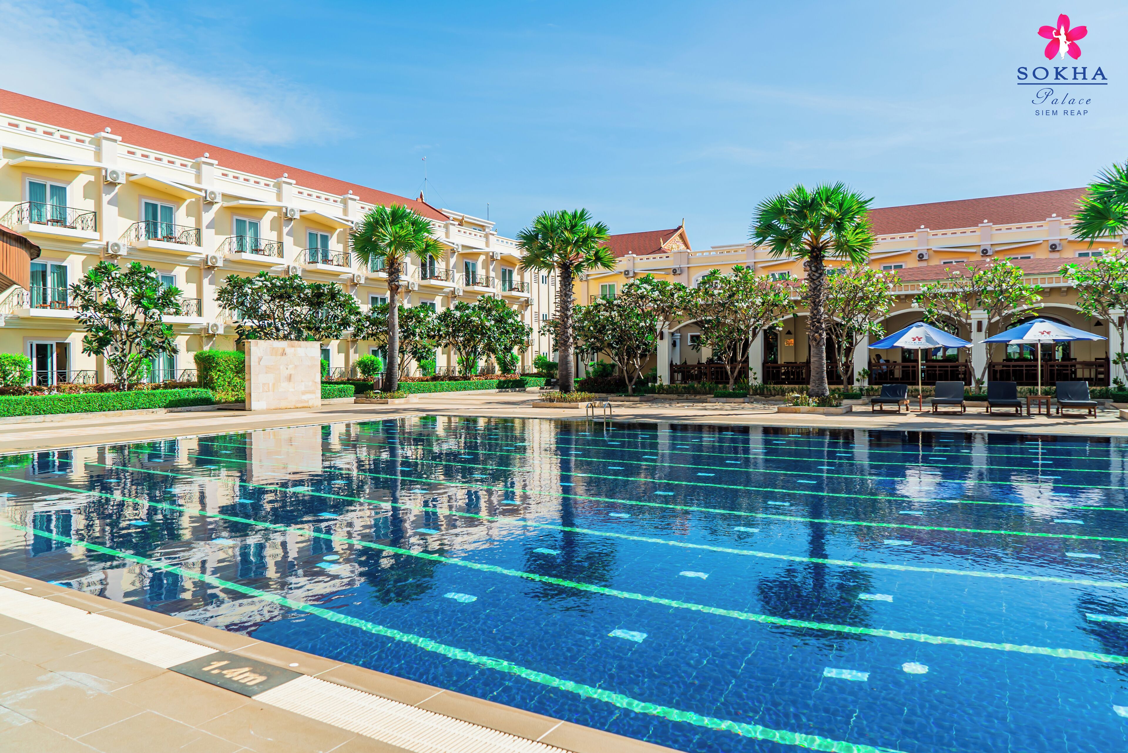 Una piscina con agua cristalina azul, rodeada por un hotel con varios balcones y palmeras.