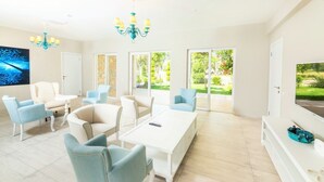 Un salon lumineux avec un canapé blanc, des fauteuils bleus, une table basse blanche et un téléviseur fixé au mur.