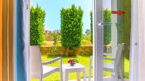 Vue depuis une pièce dotée d’une porte vitrée, avec une petite table sur laquelle est posé un vase de fleurs, deux chaises blanches et, au fond, un mur en pierre.
