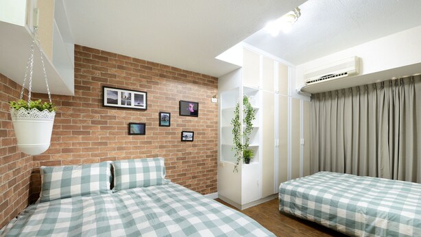 Een slaapkamer met een bakstenen muur, twee bedden met geruit beddengoed, een hangende plant en een witte airconditioning.