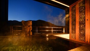 Zona de aguas termales con una escultura de piedra, una valla de madera y vistas a montañas al anochecer.