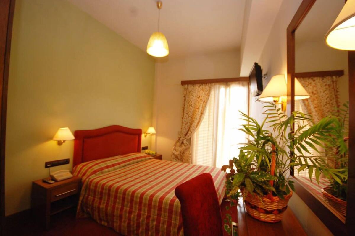 Una habitación de hotel con cama, lámparas de noche, una silla roja, un ventanal con cortinas y una planta en maceta.