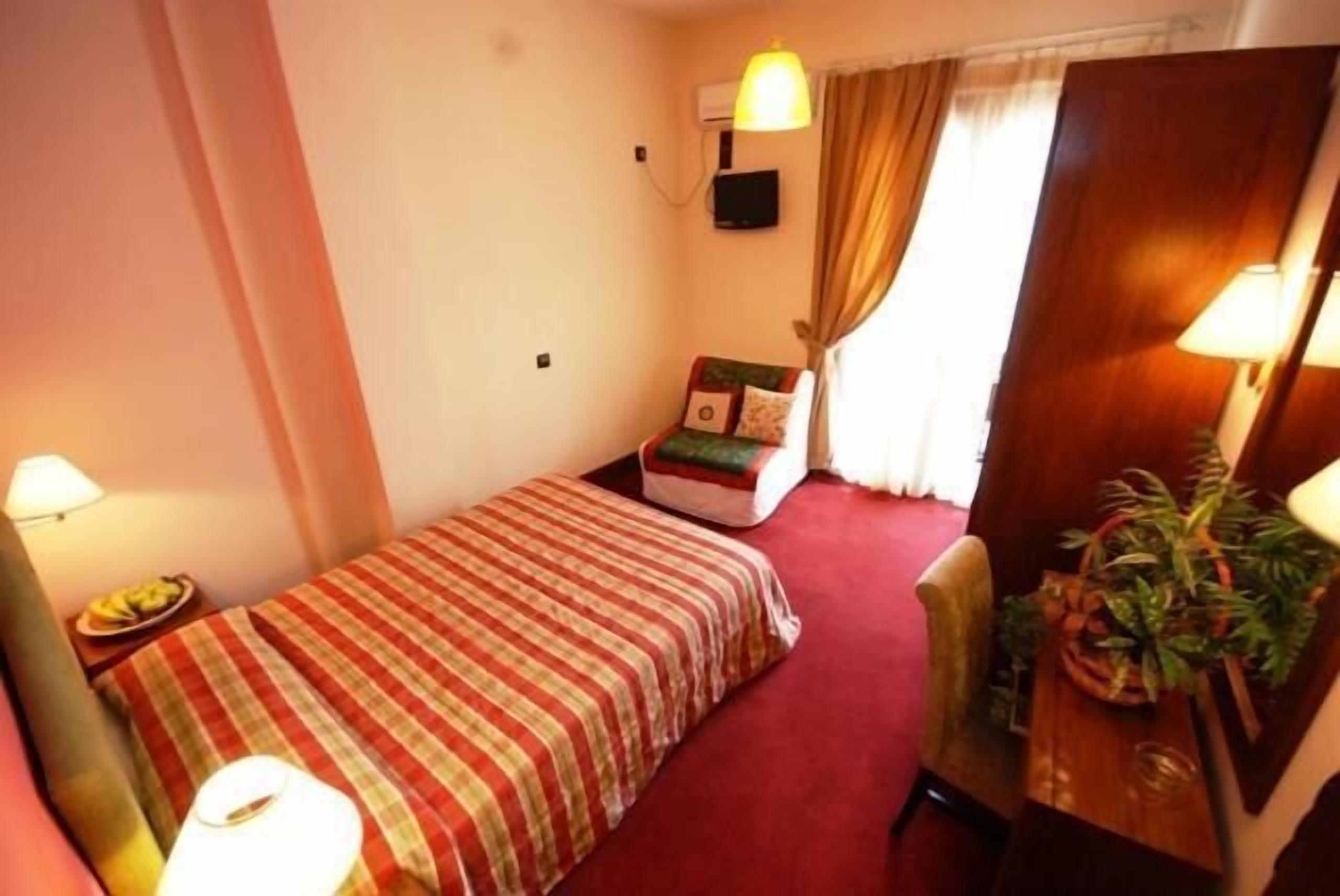 Ein kleines, gemütliches Hotelzimmer mit einem Einzelbett, einem Sessel, einem Schreibtisch mit Lampe und einem Blumentopf.