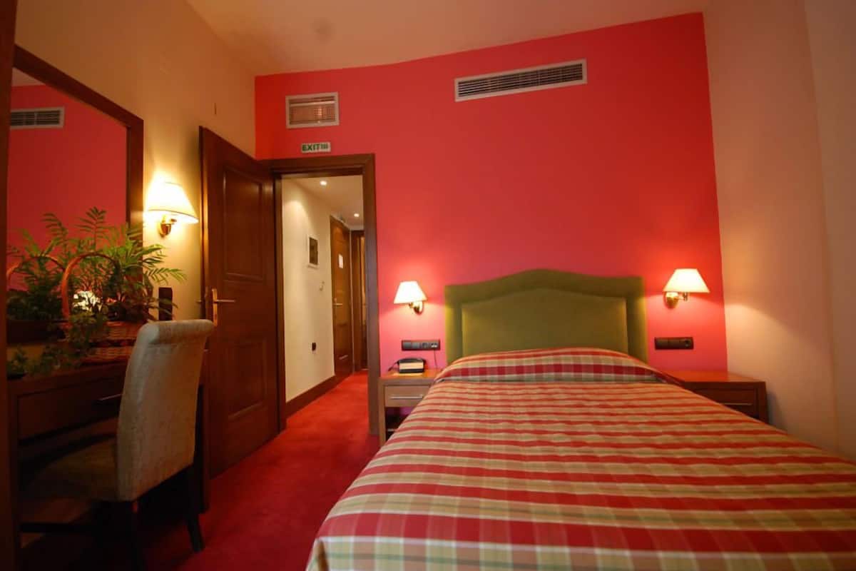 Habitación de hotel con una pared roja, un cabecero verde, colcha de cuadros, dos lámparas de noche, un escritorio con una silla y una planta en maceta.