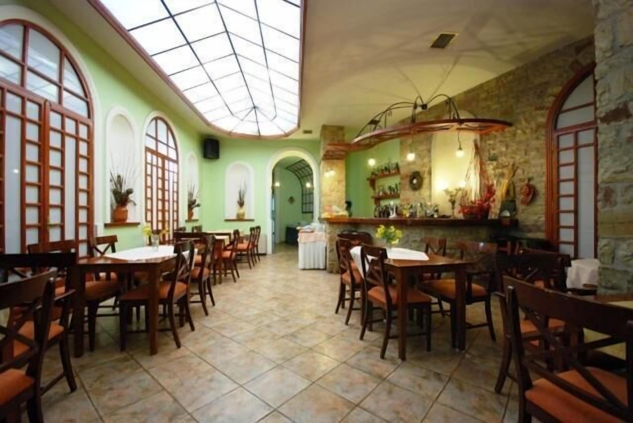 A restaurant with multiple wooden tables and chairs, a high ceiling with a skylight, and a stone wall with a bar area.