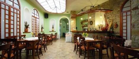 A restaurant with multiple wooden tables and chairs, a high ceiling with a skylight, and a stone wall with a bar area.