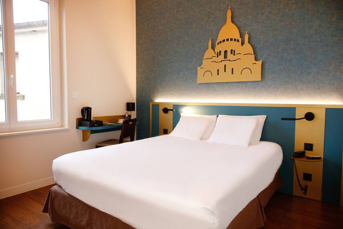 A hotel room with a large bed, a desk with a coffee maker, a wall-mounted lamp, and a decorative wall piece.