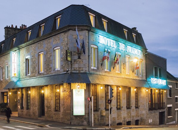 A historic stone building with a corner entrance, a restaurant sign, and flags. - Hôtel de France Vire (Vire-Normandie)